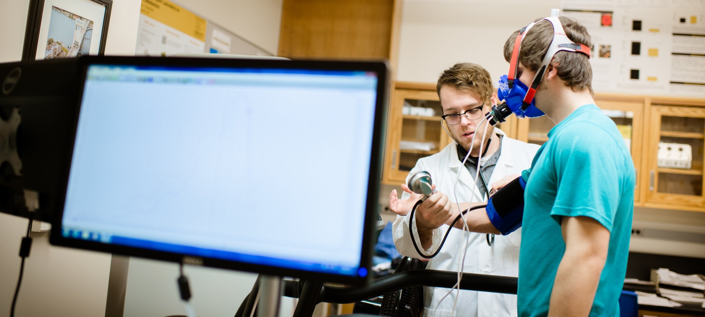 Two male students, one researcher measuring the participant's blood pressure while he is on a treadmill wearing a breathing mask