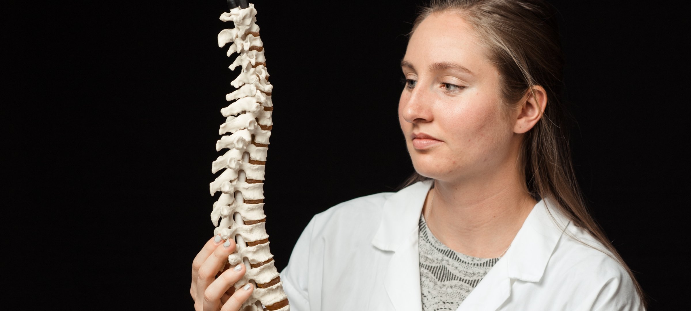 Student in a lab coat looking at a model of a spine.