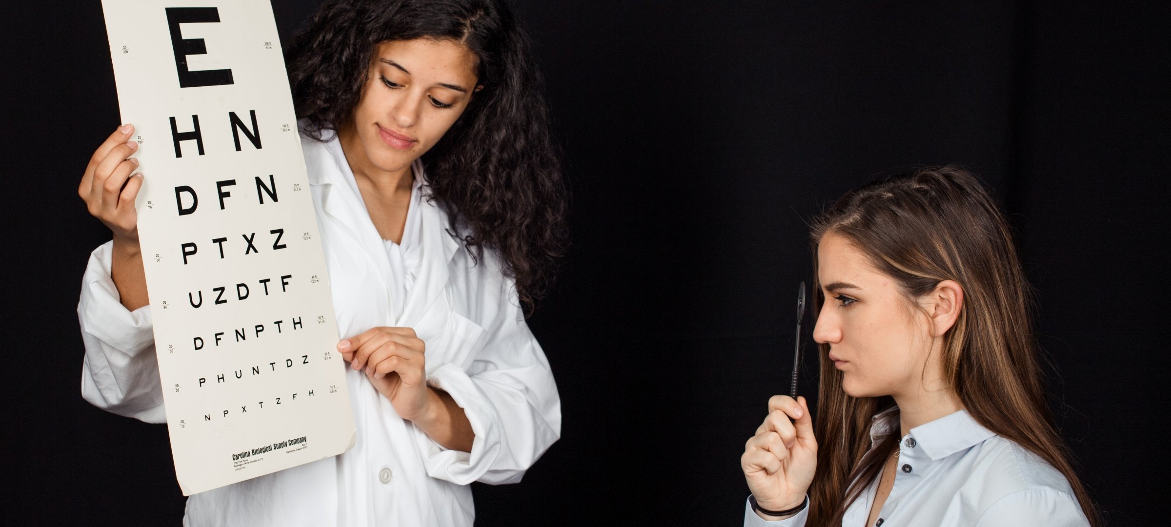 Student in a lab coat holding eye lettered chart while another holds a ophthalmoscope to the eye.