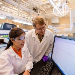 Two researchers, one male and one female, in a lab viewing results on a screen wearing lab coats, safety glasses, and gloves