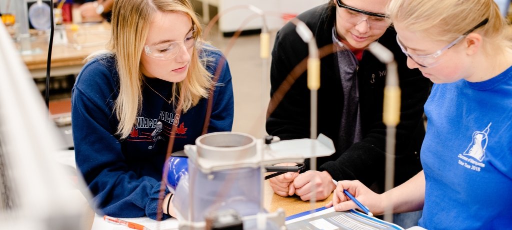 Three researchers with goggles in a lab looking over a data sheet.