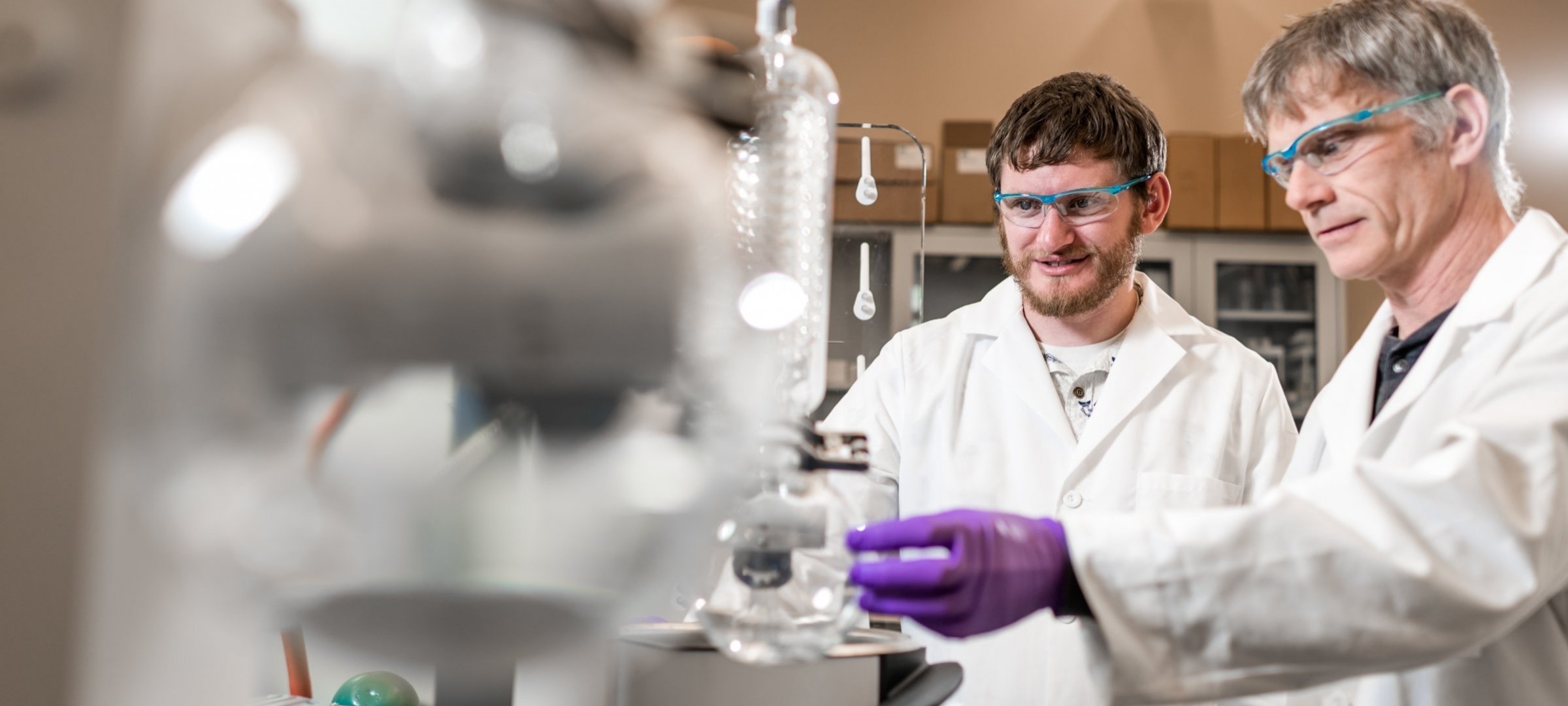 Two researchers in safety glasses, gloves and lab coats in a lab with one holding a solution