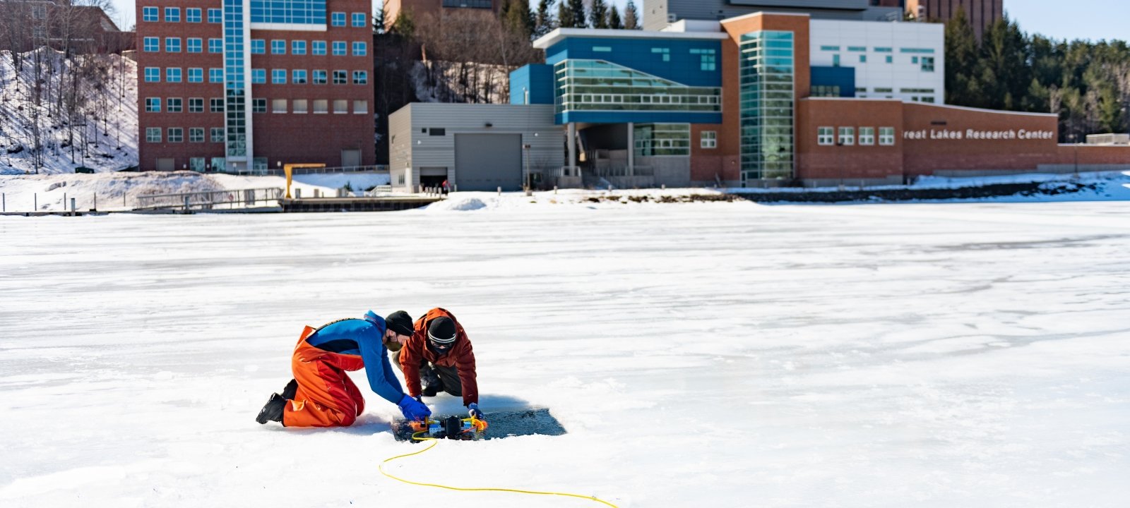 Researchers on the frozen canal across from campus dropping research equipment in the water.