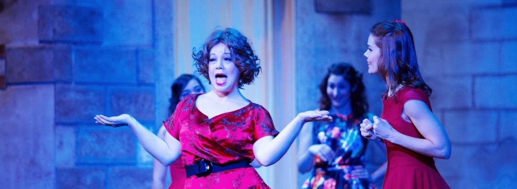 A performer stands with hands outstretched, singing, while others look on.