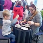 A mother having tea with her young son while he dips the tea bag into water.