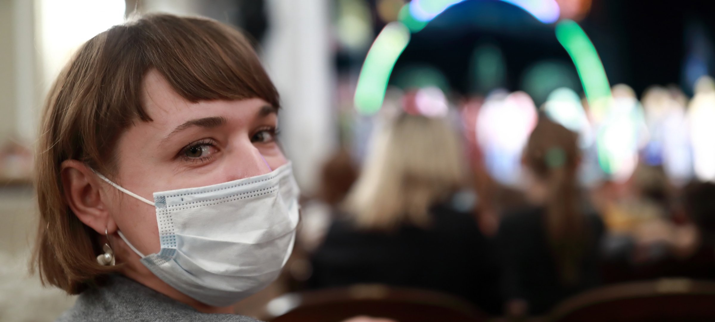 Masked person in theater audience looks over their right shoulder at the camera