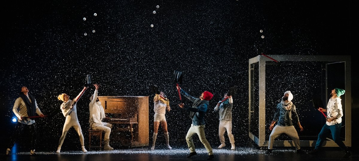 Acrobats wearing winter jackets and hats toss snowballs into the air on a snowy stage