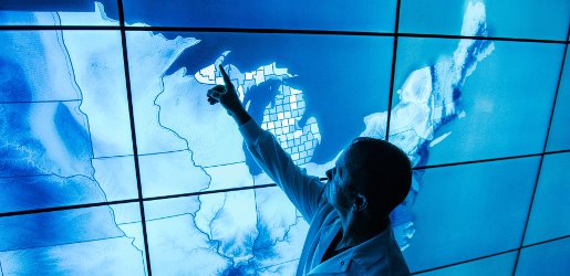 Someone pointing to Houghton, MI on a map of the northeast U.S. on a large video screen.