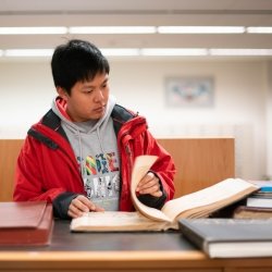 Student standing looking through a large book in the library