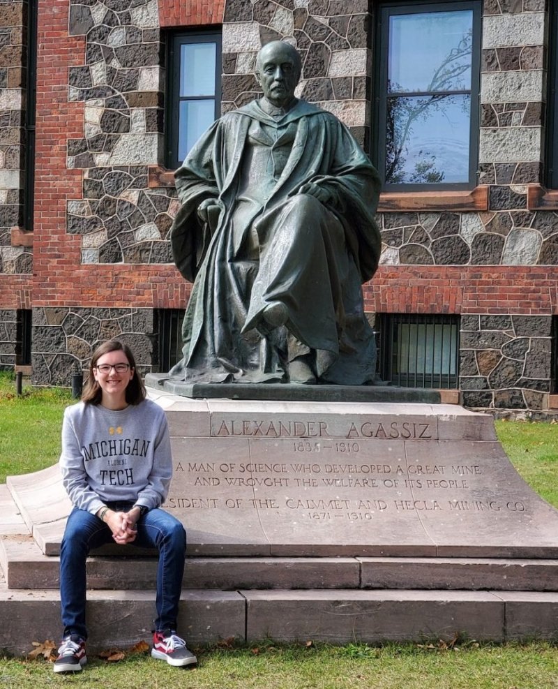 A young woman in a Michigan Tech sweatshirt sits by a statue of Alexander Agassiz in Calumet Michigan outside