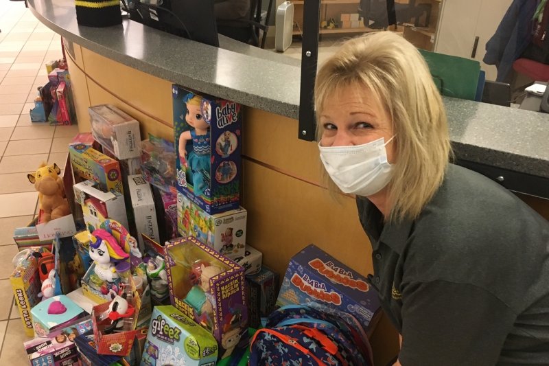 A smiling woman wears a mask and a Michigan Tech polo shirt at the front of a Library desk where the toys she has collected are piled.