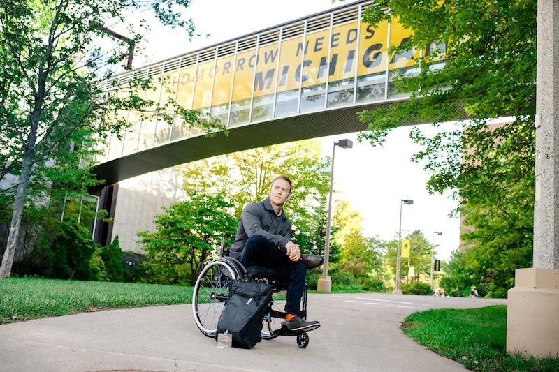 A young man sitting in a wheelchair with his backpack on a sidewalk with Tomorrow Needs Michigan Tech on the banner behind him