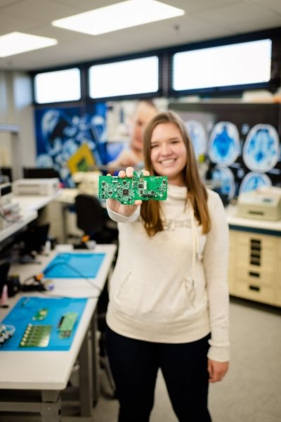 A young woman holds up a green circuit board in an electronics lab for students. Her shirt says Michigan Tech.
