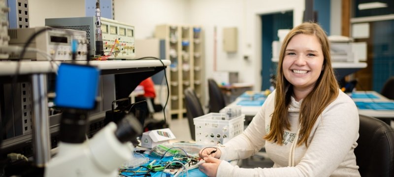 A young woman wearing a Michigan Tech shirt sits at a circuit board in an electronics makerspace at Michigan Tech with electronic equipment behind her.