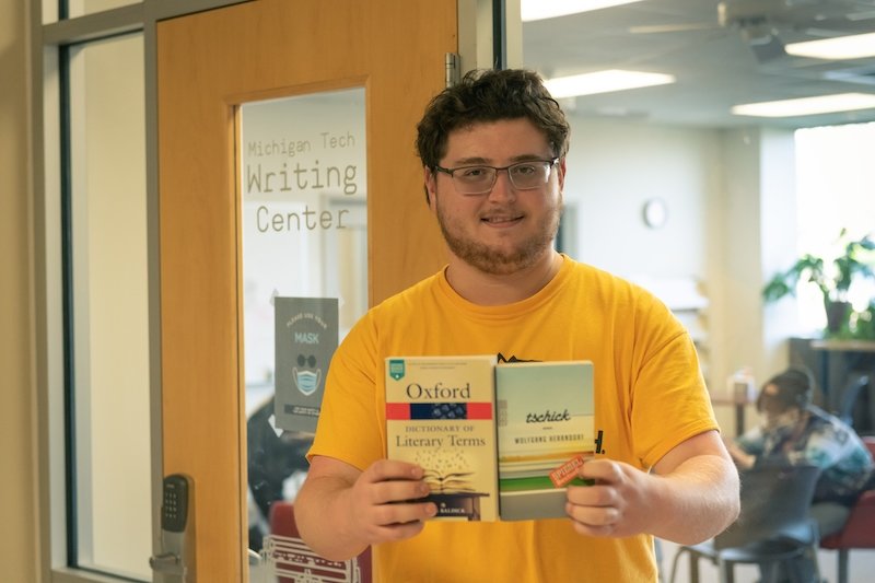 A young man holds Oxford dictionary of literary terms and a German book in his hands behind a glass door that says Michigan Tech Writing Center