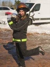 A young woman in emergency response gear and mudboots playfully swings a shovel in front of the ambulance.