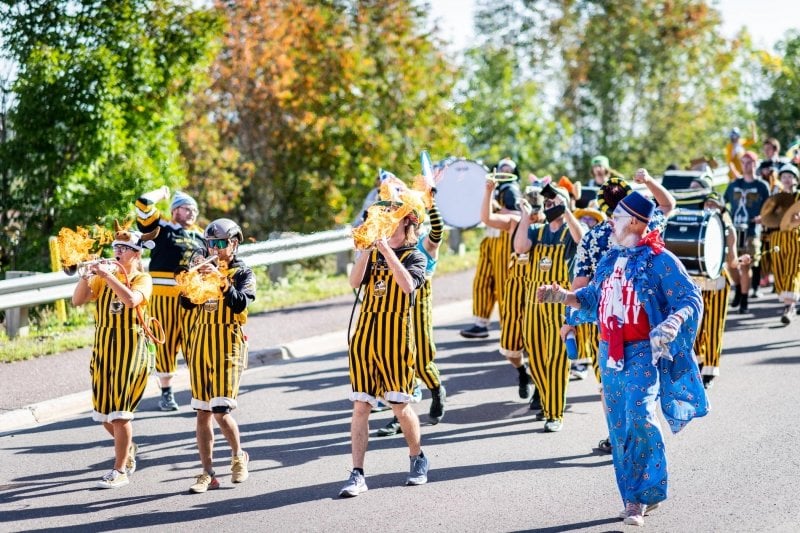 Huskies Pep Band members march with flaming trumpets as a clown walks alongside outdoors in the 2021 Parade of Nations