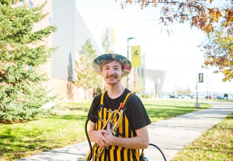 A Pep Band member from the Michigan Tech Huskies poses outside Walker Arts and Humanities Center in his striped bibs and funny hat holding his flaming trumpet on a sunny fall day.