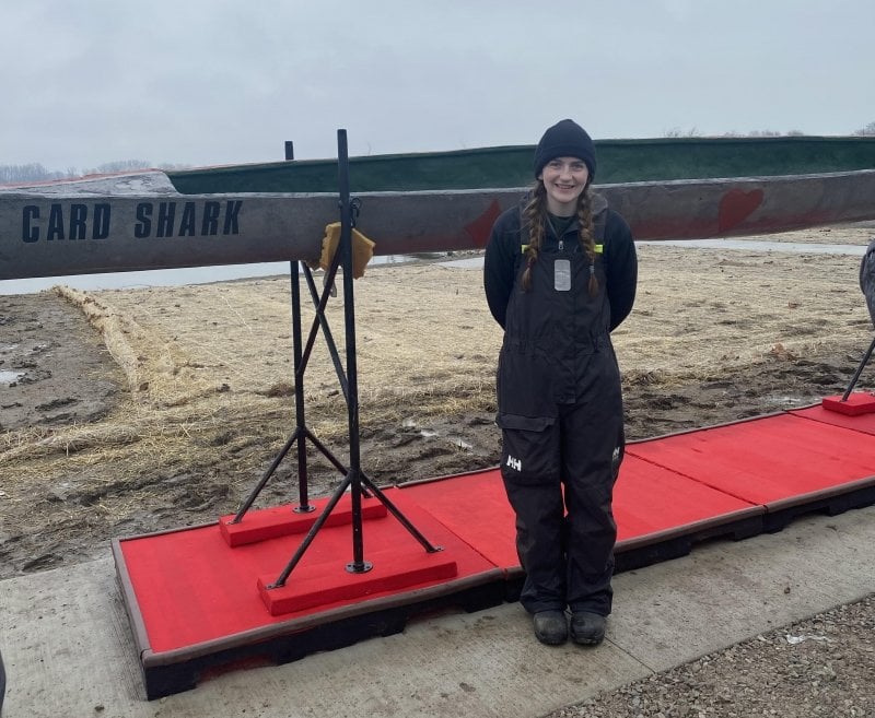 A team captain with braids in waterproof bibs, boots, and hats stands in front of Michigan Tech's concrete canoe entry. She's team co-captain.