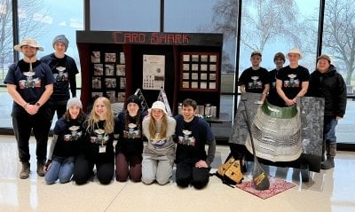 Students kneel in front of their poster presentation for Card Shark at the Concrete Canoe Regionals, while other members of the Michigan Tech team stand on each side of the presentation which includes playing cards and a sheet showing the construction materials used in the canoe.