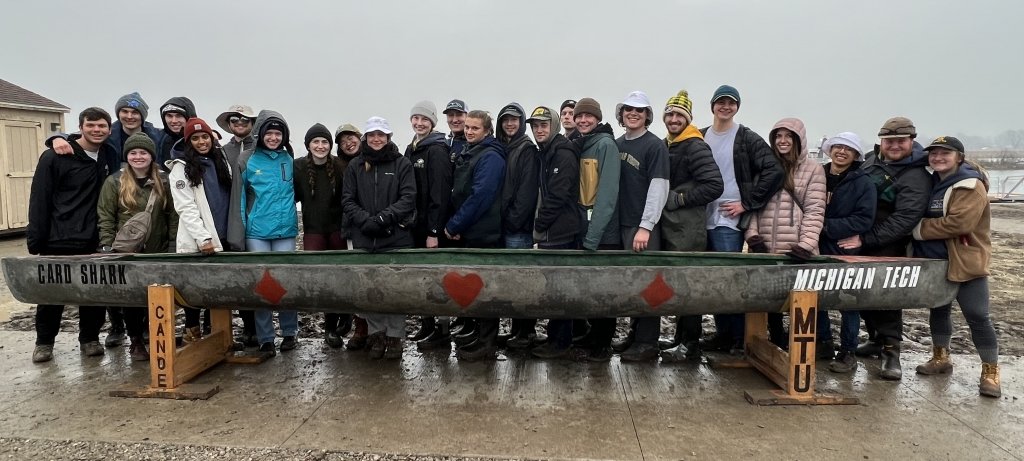 24 members of Michigan Tech Concrete Canoe Team stand behind their canoe Card Shark in a competition at a pond near Wayne State University.