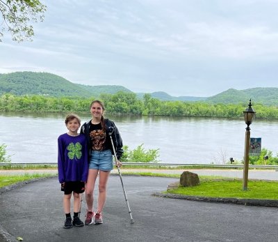 A young brother with a clover on his sweatshirt and his sister, with crutches, in front of a body of water with hills behind them at home in Wisconsin as the Michigan Tech student-athlete prepares to compete for the world's fastest running time on crutches.