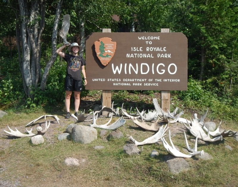 A young researcher stands by the Isle Royale Windigo sign surrouned by moose antlers and holding one up to her head. She's national scholarship winner Quinn Riordan. The Obama-Cheskey Voyager Scholar goes to school at Michigan Tech — and learned about the college when she met researcher Rolf Peterson at Isle Royale.
