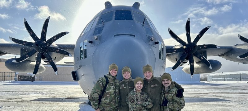 Five Air Force ROTC cadets pose in front of the nose of a C-130 airplane.