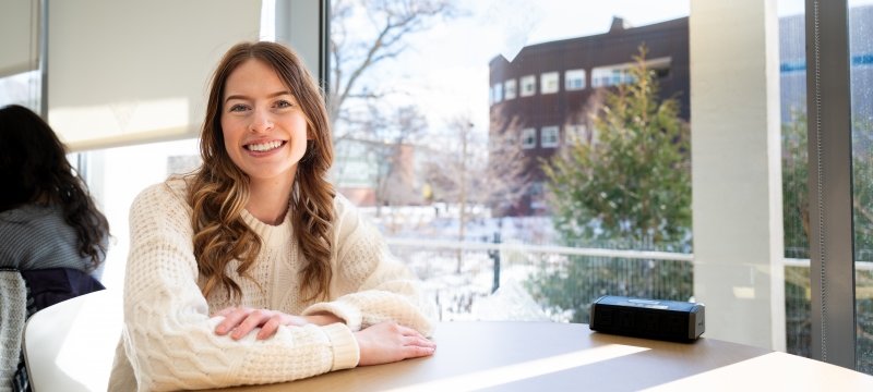 A computer science and psychology student smiles from her table in the Library with Michigan Tech's College of Computing in the background.