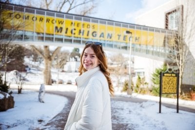 A Michigan Tech student studying computer science and psychology smiles on the snowy Michigan Tech campus with her hands in her pockets in front of the Tomorrow Needs Michigan Tech Library Walkway.