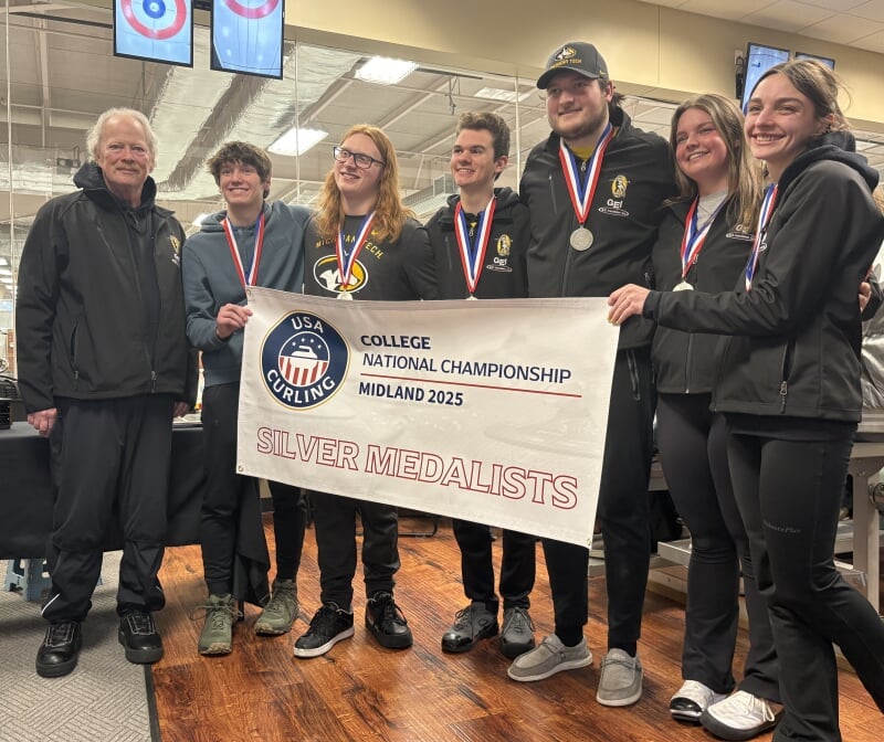 Seven smiling people who are members of a curling club at Michigan Tech stand in the winner’s circle holding a banner that reads College National Championship, Midland 2025, Silver Medalists.