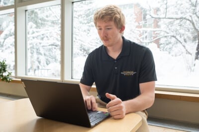 Connor Tokie works on a laptop in the Dow Building lobby, wearing a black polo that reads Steel Bridge Team at Michigan Tech.