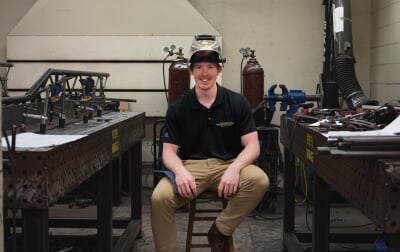 Connor Tokie in the Dow fabrication shop wearing a welding helmet and a black polo that reads Steel Bridge Team at Michigan Tech. On the table next to him is a steel beam that reads Steel Bridge.