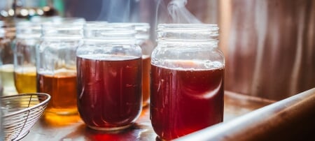 A row of glass jars filled with maple sap and syrup ranging in a gradient from clear, in the distance, to much darker closer to the camera, sit on a table in the Nara Family Maple Center. A wisp of steam rises from the two closest jars.