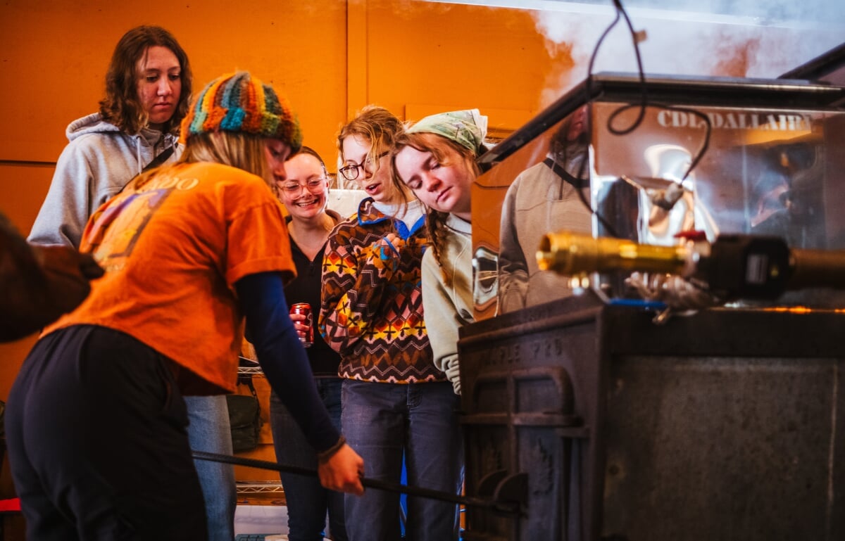 A student stokes the fire of a large boiler while four other students watch.