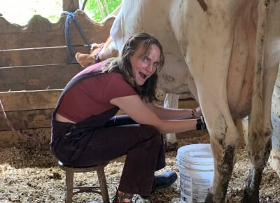 Allyson McQuiston sits on a stool in a barn milking a white and red cow.