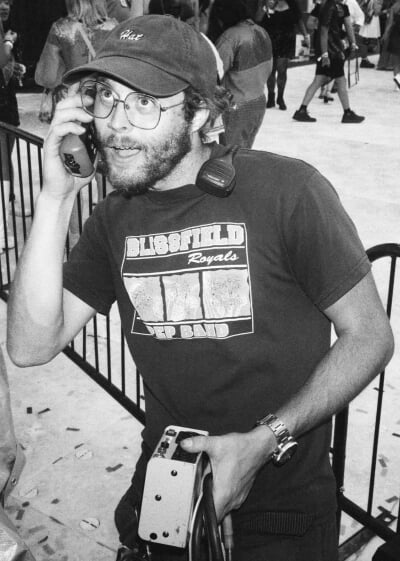 A black-and-white photo of Charles Southward at a live event, holding a piece of sound equipment to his ear like a phone as he makes a comedic expression.