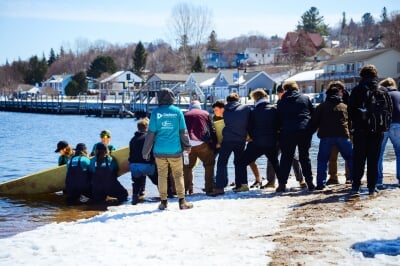 A dozen students work together to portage a large yellow concrete canoe into the water on a snowy shore.