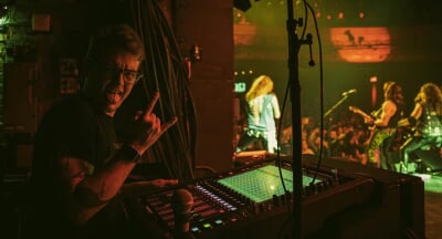 Dave Peterson stands behind a sound board backstage at a live concert, holding one hand up in a rock-and-roll gesture and sticking out his tongue.