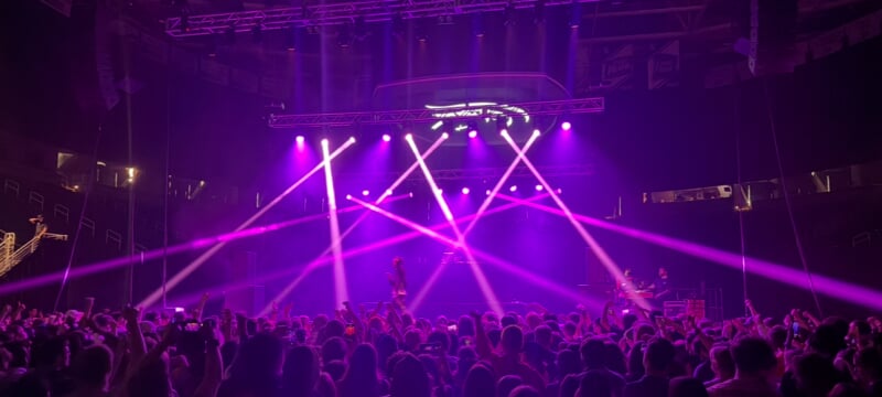 A concert venue filled with people and lit with bright purple beams from multiple stage lights. 