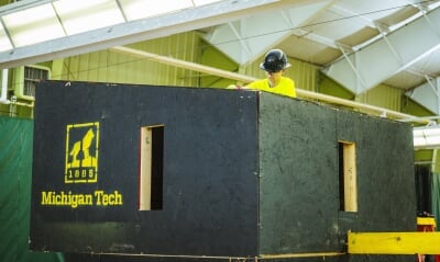 A Michigan Tech student assembles the roof of a wood building, which has been painted black with a stenciled Michigan Tech logo.