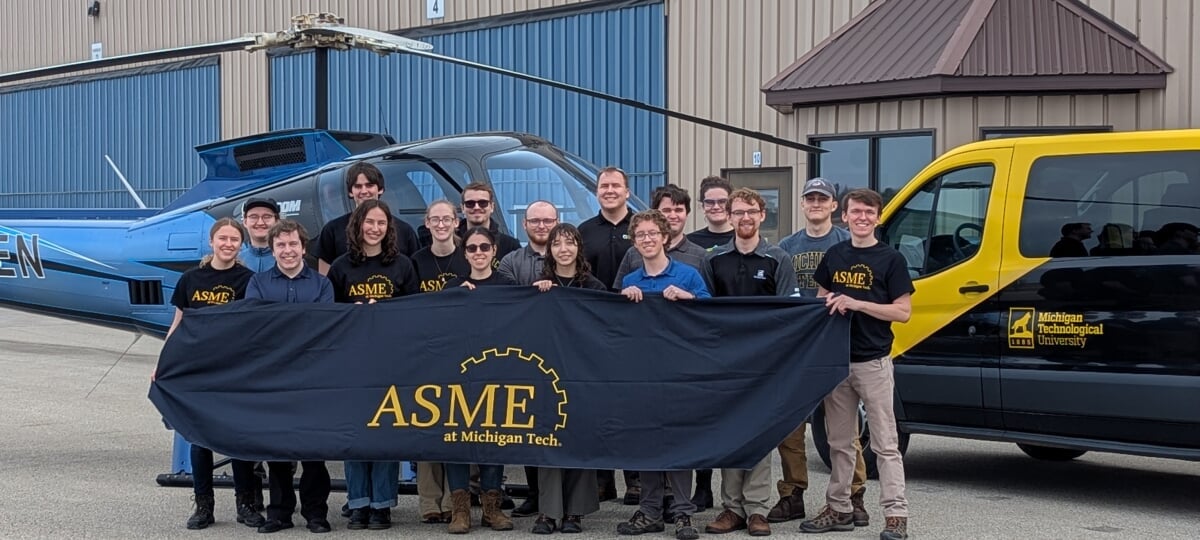 ASME students pose between an Enstrom helicopter and Michigan Tech van holding an 'ASME at Michigan Tech' banner.