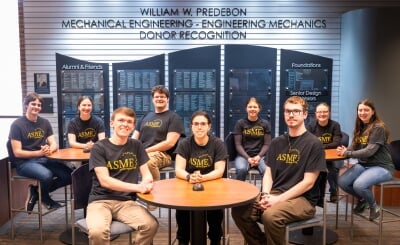 ASME members face the camera for a group photo in the lobby of the R. L. Smith Building, all wearing black ASME T-shirts and jeans, seated at three round tables