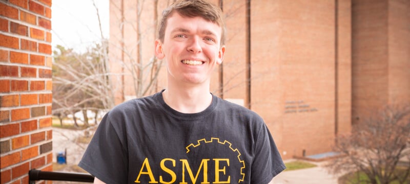 Jacob Bruesch smiles in a black ASME T-shirt on an open balcony opposite the R. L. Smith Building.