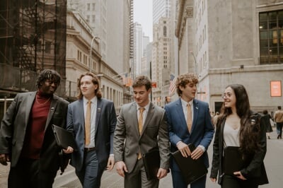 Five students in suits walking down a sidewalk with tall buildings behind them.