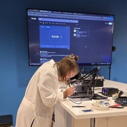 Elise Buzzell, in lab coat, works with a soldering iron on a circuit board in front of a blue screen.