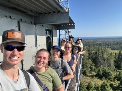Michigan Tech students smile for a photo from the top of Ojibway Tower with views of woodlands and Lake Superior in the background.