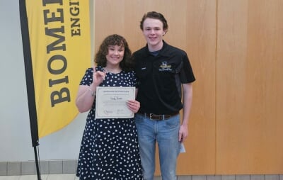 Emily Breton, holding a certificate with her right pinky raised with her Order of the Engineer ring, standing next to Jared Ott in front of a Biomedical Engineering banner.