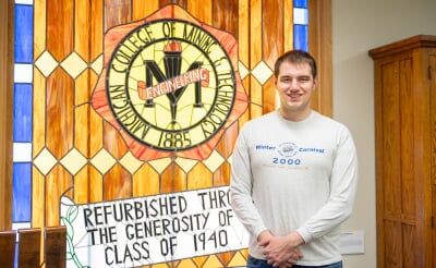 Ethan Rice, wearing a Winter Carnival 2000 shirt, stands in front of a stained glass window of an old Michigan College of Mining and Technology logo.