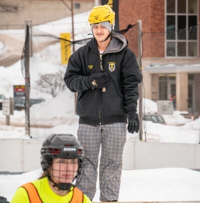 Hieu wearing a helmet and standing on a snowbank looking over a broomball rink.